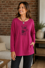 Load image into Gallery viewer, A woman wearing a bright cerise pink long-sleeve tunic top with a vintage-style graphic print on the front, paired with black pants, standing in a cozy living room setting with a brick wall background.
