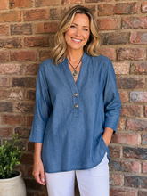 Load image into Gallery viewer, A smiling woman with blonde hair stands against a red-brown brick wall wearing a blue button-front top and white pants.
