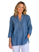 Load image into Gallery viewer, A smiling woman with blonde hair stands against a white background a blue button-front top and white pants.
