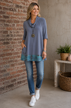Load image into Gallery viewer, A woman modeling a grey cowl-neck tunic top with a teal patterned hem, paired with blue jeans and white sneakers, standing in front of a brick wall.
