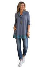 Load image into Gallery viewer, A woman modeling a grey cowl-neck tunic top with a teal patterned hem, paired with blue jeans and white sneakers, on a solid white background.
