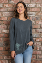 Load image into Gallery viewer, A woman wearing a dark gray long-sleeve top with a white line art graphic of a windmill and a small sign on the lower left front, paired with light blue jeans, standing against a rustic red-brown brick wall.
