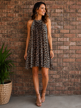 Load image into Gallery viewer, A woman wearing a sleeveless leopard-print mini dress stands in front of a brick wall, smiling and looking to the side. She is wearing tan heeled sandals, and a potted plant is visible on the left.
