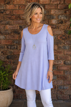 Load image into Gallery viewer, A woman wearing a light lavender cold-shoulder tunic top and white pants, standing in front of a rustic brick wall.
