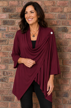 Load image into Gallery viewer, A woman wearing a draped burgundy wrap top over a black top and black pants, standing against a red-brown brick wall.
