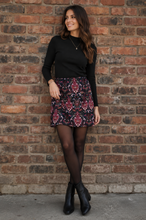 Load image into Gallery viewer, A woman stands against a red-brown brick wall wearing a black long-sleeve top, a patterned mini skirt with red, blue, and white motifs, sheer black tights, and black ankle boots.
