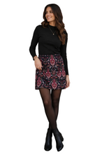Load image into Gallery viewer, A woman stands against a red-brown brick wall wearing a black long-sleeve top, a patterned mini skirt with red, blue, and white motifs, sheer black tights, and black ankle boots.

