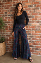 Load image into Gallery viewer, A woman stands against a red brick wall wearing a black long-sleeve scoop-neck top and high-waisted, wide-leg pants with a dark blue and red geometric pattern. She is wearing black flip-flop sandals and standing next to a potted plant.
