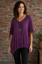 Load image into Gallery viewer, A woman wearing a purple short-sleeve tunic top with tie details on the sleeves, paired with black pants, standing in front of a brick wall.
