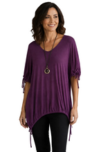 Load image into Gallery viewer, A woman wearing a purple short-sleeve tunic top with tie details on the sleeves, paired with black pants, standing in against a white background.  

