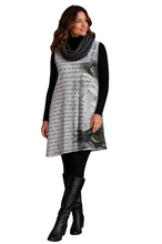 Load image into Gallery viewer, A woman standing against a white background, wearing a sleeveless black-and-white graphic tunic dress with a gray knit cowl scarf, black leggings, and black knee-high boots.
