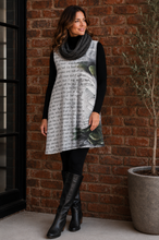 Load image into Gallery viewer, A woman stands outdoors against a red brick wall wearing a sleeveless black-and-white graphic tunic dress with a gray knit cowl scarf, black leggings, and black knee-high boots.
