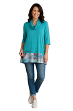 Load image into Gallery viewer, A woman stands against a white background, wearing a turquoise cowl-neck tunic top with a tie-dye patterned hem, blue jeans, and white sneakers.
