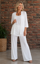 Load image into Gallery viewer, A woman modeling a white outfit consisting of a short-sleeve open-front cardigan, a scoop-neck top, hip skirt and matching pants, standing in front of a brick wall.

