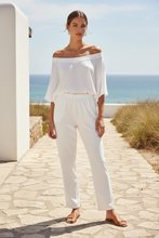 Load image into Gallery viewer, A woman modeling a white off-the-shoulder top and matching white pants on a stone path with a beach and ocean in the background.
