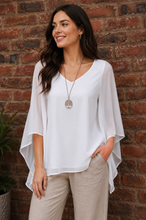 Load image into Gallery viewer, A woman wearing a white V-neck blouse with wide, draped batwing sleeves and light beige pants, standing against a red brick wall.
