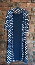 Load image into Gallery viewer, A navy blue open-front cardigan with white polka dots layered over a solid navy blue dress, hanging on a grey hanger against a red-brown brick wall.
