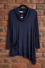 Load image into Gallery viewer, A navy blue long-sleeve cowl neck top with asymmetrical hemline, hanging on a black hanger against a red-brown brick wall.
