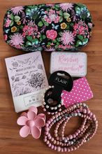 Load image into Gallery viewer, A flat-lay collection of various beauty and lifestyle accessories on a wooden surface, including a floral makeup bag, a pink flower-shaped hair clip, a beaded necklace, a small mirror, ponytail hair bands, a small tin with floral text, and a notebook with botanical illustrations.
