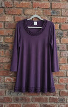 Load image into Gallery viewer, A purple long-sleeve tunic top with ruffled trim at the neckline and hem, hanging on a gray hanger against a red brick wall.
