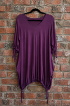 Load image into Gallery viewer, A purple long-sleeve tunic top with side tie details and a gathered drawstring hem, hanging on a black hanger against a red brick wall.
