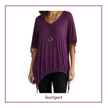 Load image into Gallery viewer, A woman wearing a purple short-sleeve top with tie details at the cuffs and hem, paired with black pants and a long necklace, shown against a white background with a purple border. The brand name 'hartgoet' is printed at the bottom.
