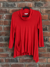 Load image into Gallery viewer, A red longsleeve top with a cowl neck and asymmetrical hemline on a hanger, hanging against a brick wall.
