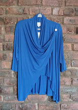 Load image into Gallery viewer, A bright blue draped wrap-style top hanging on a brick wall. The top features a draped neckline, three-quarter sleeves, and an asymmetrical draped hem, with a small white decorative button on the upper right side.
