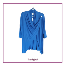 Load image into Gallery viewer, A bright blue draped wrap-style top with three-quarter sleeves and an asymmetrical hem, hanging on a hanger against a white background with a purple border and the brand name hartgoet below.
