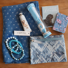 Load image into Gallery viewer, A curated flat-lay of blue-themed items on a wooden surface, including a blue polka-dot scarf, a mini manicure set in a clear case, a small handcream, a small handbag mirror, small notebook, a patterned zip pouch, and 3 beaded bracelets.
