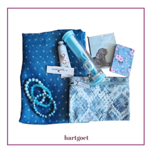 Load image into Gallery viewer, A flat-lay of a blue gift set on a white background with a purple border. The set includes a blue polka-dot scarf, a blue patterned zippered pouch, a light blue tube, 3 blue beaded bracelets, a small handcream, small mirror and a small notebook.

