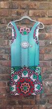 Load image into Gallery viewer, A sleeveless dress with colorful mandala patterns in pink, red, green, and white, hanging on a gray hanger against a rustic red brick wall.
