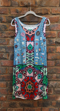 Load image into Gallery viewer, A sleeveless bohemian-style dress hanging on a white plastic hanger against a rustic red-brown brick wall. The dress features a light blue denim-look top section decorated with colorful patches, and a pink, red, green, and blue geometric and floral mandala on the bottom section.  
