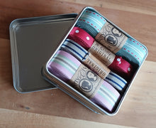 Load image into Gallery viewer, A square metal tin filled with four rolls of decorative ribbon in assorted colors and patterns, with the lid resting beside the tin on a wooden surface.
