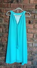 Load image into Gallery viewer, Back view of a sleeveless turquoise blue  dress with a tie detail,  hanging on a grey hanger against a red-brown brick wall.
