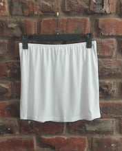 Load image into Gallery viewer, A white hip skirt hanging from a black hanger against a red brick wall background.

