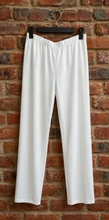 Load image into Gallery viewer, White pants hanging from a black hanger against a red-brown brick wall.
