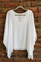 Load image into Gallery viewer, A white draped blouse with a wide scoop neckline, hanging on a gray hanger against a red-brown brick wall.
