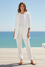 Load image into Gallery viewer, A woman stands on a wooden deck with the ocean and blue sky behind her, wearing a white coordinated outfit consisting of a sleeveless open-front cardigan, white top, white pants, and brown flat sandals.
