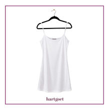 Load image into Gallery viewer, A white spaghetti-strap camisole top hanging on a black hanger against a white background, framed by a purple border with the brand name hartgoet printed below.
